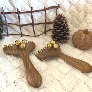 以下が含まれることがあります： 白いニットの表面に、金色の球が付いた2つの木製マッサージツール。松ぼっくり、小さな織りのカボチャ、装飾的な木製のフェンスも存在します。ツールは湾曲したハンドルを持っています。
