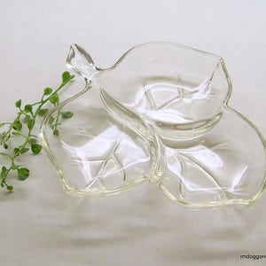 May include: A clear glass serving dish shaped like three leaves. The dish is designed for serving snacks or appetizers.