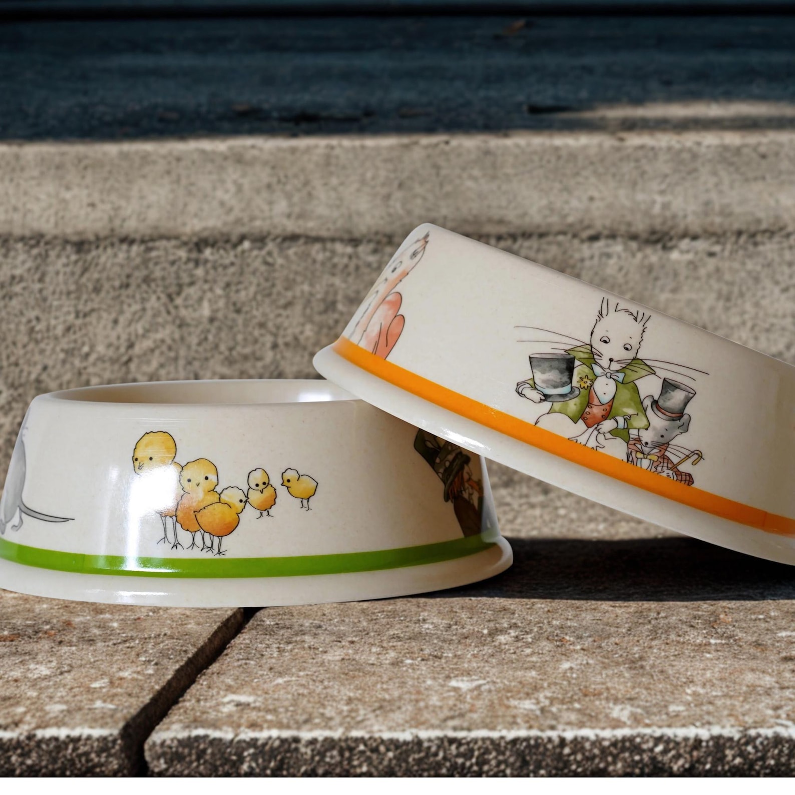 Two ceramic pet bowls are depicted, one with illustrations of chicks and a green rim, and the other with a rabbit and mouse in fancy attire with an orange rim.