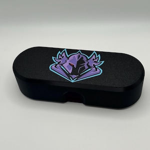 May include: A black rectangular storage case with rounded ends featuring a stylized purple and light blue graphic of a helmeted figure with red eyes centered on the top lid. The case has a smooth matte finish and a small cutout on the front edge.