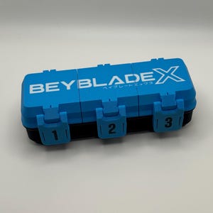 May include: A rectangular storage case with a black base and a bright blue lid featuring the white text BEYBLADEX. The lid has three separate compartments, each secured by a blue latch marked with the black numbers 1, 2, and 3 respectively.