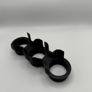 May include: A black, three-finger ring, likely 3D-printed, with three circular finger holes connected by a solid structure. The ring has a matte finish and is set against a plain white background. The design features angular shapes and a utilitarian aesthetic.