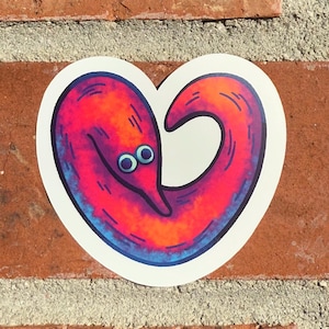 May include: A heart-shaped sticker featuring a cartoon worm with a smiling face. The worm is primarily red and orange, with blue and purple accents. The sticker is on a brick wall.