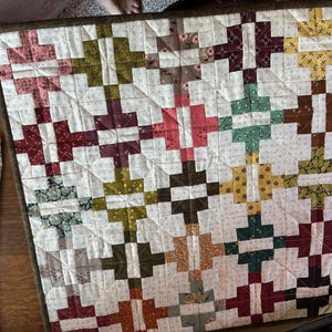 May include: A handmade quilt with a repeating pattern of colorful blocks. The blocks are arranged in a diagonal pattern, with colors including red, green, yellow, and brown. The quilt is bordered with a dark brown fabric.