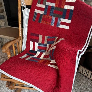 May include: A red quilt with a patchwork design featuring rectangles of various colors, including red, blue, and white. The quilt is draped over a wooden rocking chair. The quilt has a textured surface and a white border.