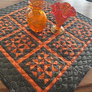 May include: An orange glass pitcher and a ruffled vase sit on a quilted table runner. The runner features a black background with orange geometric shapes and borders. The pitcher has a crackle finish.
