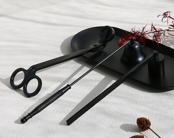 May include: A black candle care set comprising a wick trimmer, a wick dipper, and a snuffer, presented on a black tray. The wick trimmer resembles scissors with large finger holes. The set is for candle maintenance.