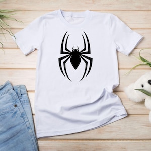 May include: White t-shirt featuring a large black spider graphic on the front. The spider design is a stylized silhouette with eight legs and a detailed body. The shirt is displayed with blue jeans and a stuffed animal.