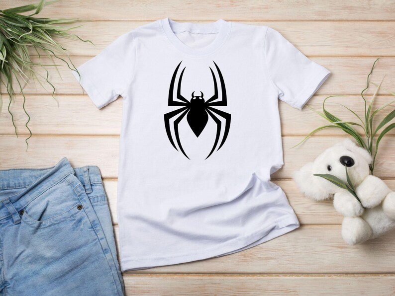 May include: White t-shirt featuring a large black spider graphic on the front. The spider design is a stylized silhouette with eight legs and a detailed body. The shirt is displayed with blue jeans and a stuffed animal.