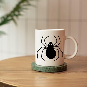 May include: White ceramic mug with a black spider graphic. The mug sits on a green marble coaster on a wooden table. The spider design features a black body and thin, curved legs.