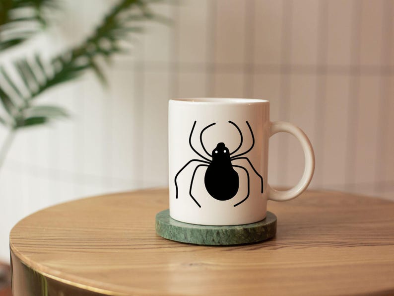 May include: White ceramic mug with a black spider graphic. The mug sits on a green marble coaster on a wooden table. The spider design features a black body and thin, curved legs.