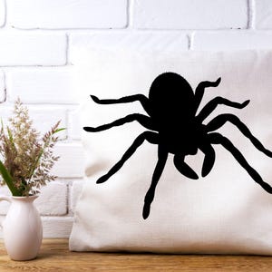 May include: A white square pillow with a large black spider silhouette. The spider has eight legs and a round body. The pillow is on a wooden surface, with a white brick wall in the background.