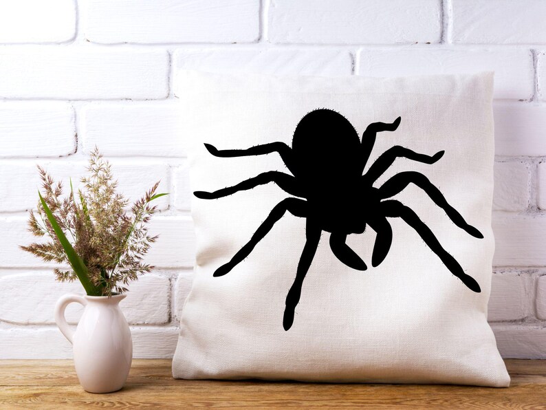 May include: A white square pillow with a large black spider silhouette. The spider has eight legs and a round body. The pillow is on a wooden surface, with a white brick wall in the background.