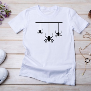 May include: White t-shirt featuring a black graphic of three spiders hanging from a horizontal line. The shirt is displayed on a light wood surface, with blue sneakers, sunglasses, and jewelry nearby. A purple flower arrangement is also present.