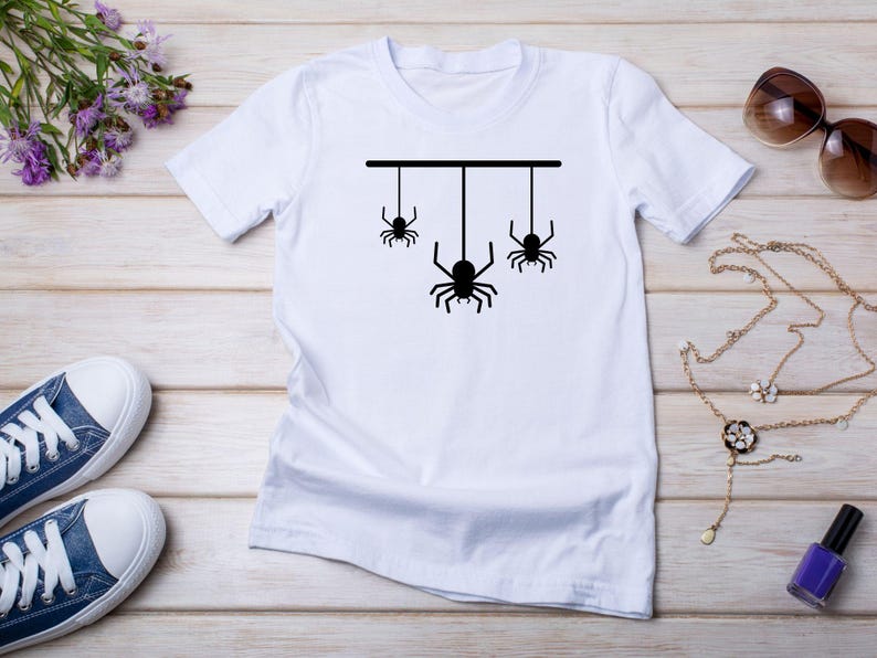 May include: White t-shirt featuring a black graphic of three spiders hanging from a horizontal line. The shirt is displayed on a light wood surface, with blue sneakers, sunglasses, and jewelry nearby. A purple flower arrangement is also present.