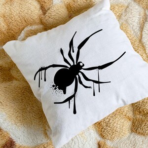 May include: White square pillow featuring a black spider design. The spider has a round body and long legs, with dripping paint effect. The pillow is placed on a textured, tan and brown surface.