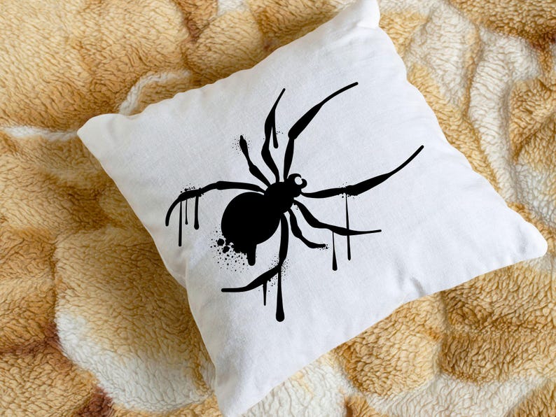 May include: White square pillow featuring a black spider design. The spider has a round body and long legs, with dripping paint effect. The pillow is placed on a textured, tan and brown surface.