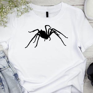 May include: White t-shirt featuring a large, black spider silhouette. The spider graphic is centered on the shirt, with its legs outstretched. The shirt is made of a soft material and is suitable for casual wear.