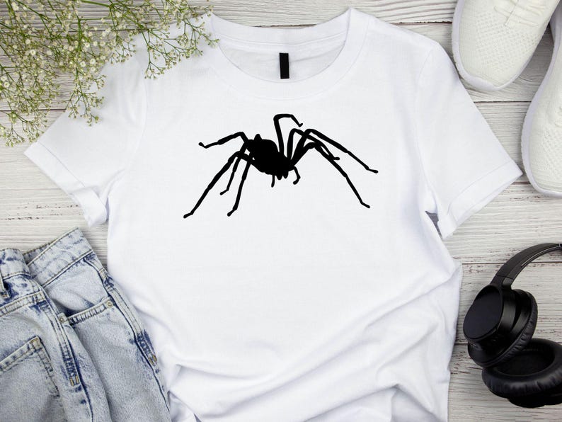 May include: White t-shirt featuring a large, black spider silhouette. The spider graphic is centered on the shirt, with its legs outstretched. The shirt is made of a soft material and is suitable for casual wear.