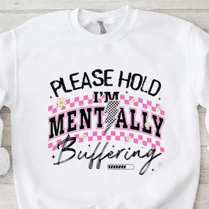 May include: White sweatshirt with the text "PLEASE HOLD I'M MENTALLY Buffering" in black and pink. The design includes a lightning bolt, daisies, and a loading bar graphic. The sweatshirt is a casual, comfortable style.