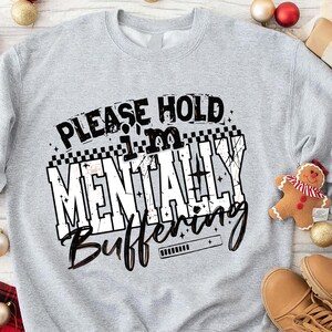 May include: A light gray sweatshirt with the text "PLEASE HOLD I'M MENTALLY BUFFERING" in a black and white design. The sweatshirt is surrounded by Christmas decorations, including ornaments, a gingerbread man, and boots.