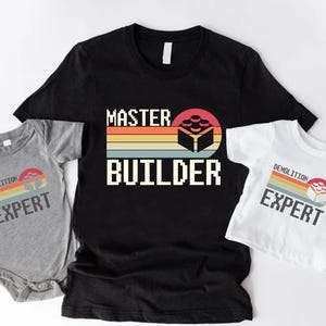 May include: A black t-shirt with "MASTER BUILDER" text and a retro graphic of a building block design. A gray onesie and a white t-shirt with "DEMOLITION EXPERT" text and a similar graphic are also displayed.