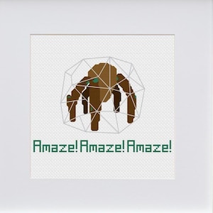Puede incluir: Una obra de arte en punto de cruz enmarcada que presenta una araña marrón dentro de una jaula geométrica. El texto verde "Amaze! Amaze! Amaze!" se muestra debajo. La obra está en un marco blanco.