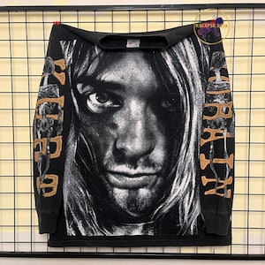 Puede incluir: Camiseta negra de manga larga con un retrato en blanco y negro. Las mangas muestran texto dorado que dice "Kurt" y "Cobain". La camiseta se muestra sobre un fondo de rejilla.