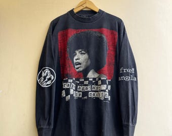 May include: A black long-sleeve t-shirt featuring a graphic of Angela Davis on a red background. Below the portrait is a checkered pattern with the text "rage against the machine". The right sleeve says "free!! angela" and the left has a circular logo.