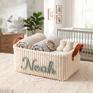 Op de afbeelding: Een crèmekleurige corduroy opbergmand met bruine leren handvatten, gevuld met opgerolde dekens, een houten bijtring en babyschoentjes. De naam "Noah" staat in groene wol op de voorkant.