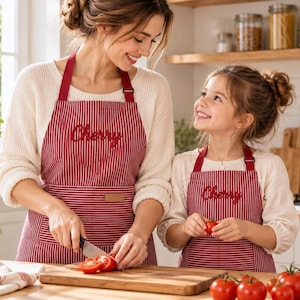 Può includere: Due grembiuli abbinati con un motivo a righe verticali rosse e bianche e la parola Cherry ricamata in corsivo rosso sul petto. Ogni grembiule include lacci rossi regolabili per il collo e una tasca frontale con una piccola etichetta rettangolare color cuoio.
