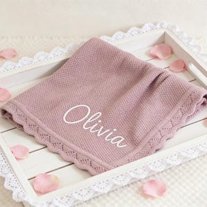 May include: A mauve-colored baby blanket with the name "Olivia" embroidered in white script. The blanket has a decorative, scalloped edge and is presented on a white tray with lace trim, scattered with pink rose petals.
