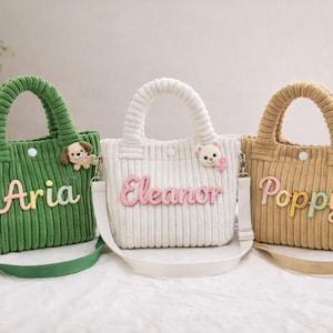 Könnte beinhalten: Drei Cord-Tragetaschen in Grün, Weiß und Beige, jede mit einem aufgestickten Namen auf der Vorderseite: "Aria", "Eleanor" und "Poppy". Die Taschen haben passende Griffe und verstellbare Schultergurte. Dekorative Tier- und Blumenakzente sind an den Taschen befestigt.