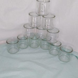 Puede incluir: Una colección de portavelas votivas de vidrio transparente dispuestas en forma de pirámide. Los soportes cilíndricos tienen una base gruesa y una abertura ancha. La disposición está sobre una superficie azul claro y blanca.