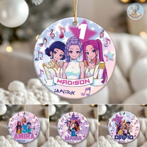 May include: A round ceramic ornament featuring three anime-style figures with purple, blue, and pink hair. The ornament has a light blue background with musical notes and stars. The name "MADISON" is displayed on a banner. The number "1" is at the top.
