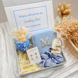May include: A blue-themed gift box with a corduroy pouch, earrings, scrunchie, and hair clips. The pouch has a pearl-studded "M". The box includes a card with the text "Megan, will you be my Something Blue?" and dried flowers.