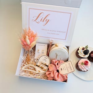 Könnte beinhalten: Eine weiße Geschenkbox mit roségoldfarbenem Rand, gefüllt mit verschiedenen Artikeln. Enthält ein Blumenarrangement, Schmuck, Haaraccessoires und eine personalisierte Nachricht. Cupcakes sind ebenfalls auf einem weißen Teller zu sehen.