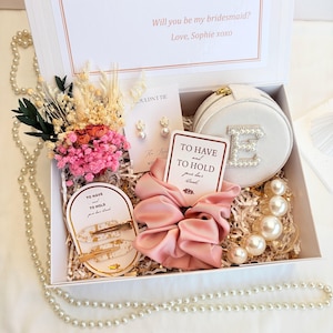 May include: A white gift box containing a round velvet jewellery case with a pearl letter E, a pink satin scrunchie, two gold-flecked hair clips, pearl earrings, a pearl hair claw, and a dried flower bouquet with the text Will you be my bridesmaid?