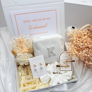 May include: A white gift box containing a corduroy pouch with a pearl letter "K", a card reading "Katie, will you be my Bridesmaid?", earrings, hair clips, and a pearl bracelet. The box is decorated with dried flowers and white ribbon, creating a cohesive theme.