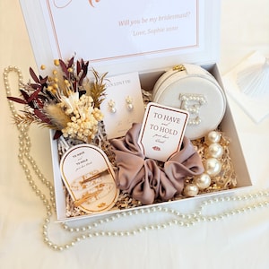 May include: A white gift box containing a dried floral bouquet, a pair of pearl earrings, a round beige jewellery case with a pearl letter E, a mauve satin scrunchie, two gold-flecked hair clips, and a string of faux pearls. Text reads Will you be my bridesmaid? Love, Sophie xoxo, To have and to hold your hair back.