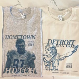 May include: Four crew neck t-shirts in cream and heather gray featuring dark blue graphic prints. The designs include portraits of football figures with text such as DETROIT DADDY, HOMETOWN HUTCH, DETROIT SUN GOD, and JUKEBOX HERO, plus 2023 2024 dates.