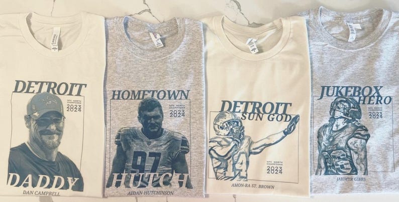May include: Four crew neck t-shirts in cream and heather gray featuring dark blue graphic prints. The designs include portraits of football figures with text such as DETROIT DADDY, HOMETOWN HUTCH, DETROIT SUN GOD, and JUKEBOX HERO, plus 2023 2024 dates.