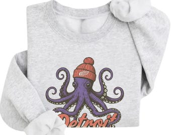 May include: A light gray crewneck sweatshirt featuring a graphic of a purple octopus wearing a red knit beanie with a white winged wheel logo. Below the octopus, the word Detroit is printed in a stylized orange font with a dark outline.