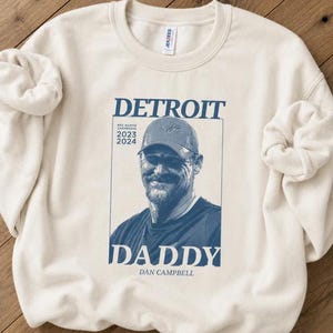 May include: A cream-colored crewneck sweatshirt featuring a blue graphic print. The design includes the text DETROIT, NFC NORTH CHAMPIONS 2023 2024, DADDY, and DAN CAMPBELL, along with a portrait of a person wearing a baseball cap in the center.