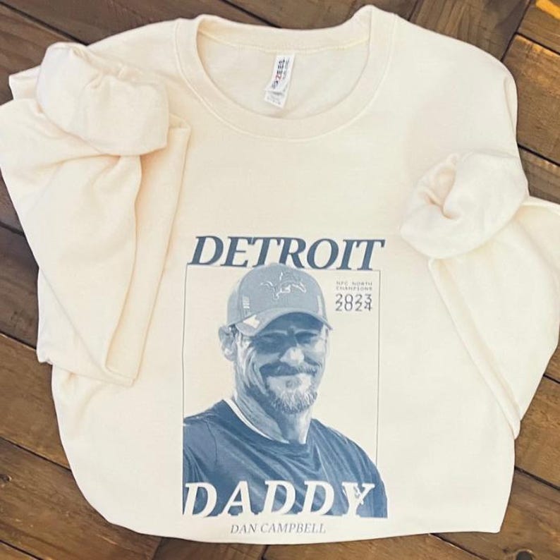 May include: A cream-colored long-sleeve sweatshirt featuring a dark blue graphic print of Dan Campbell wearing a baseball cap. The text reads DETROIT, DADDY, DAN CAMPBELL, and NFC NORTH CHAMPIONS 2023 2024. The garment has a crew neckline.