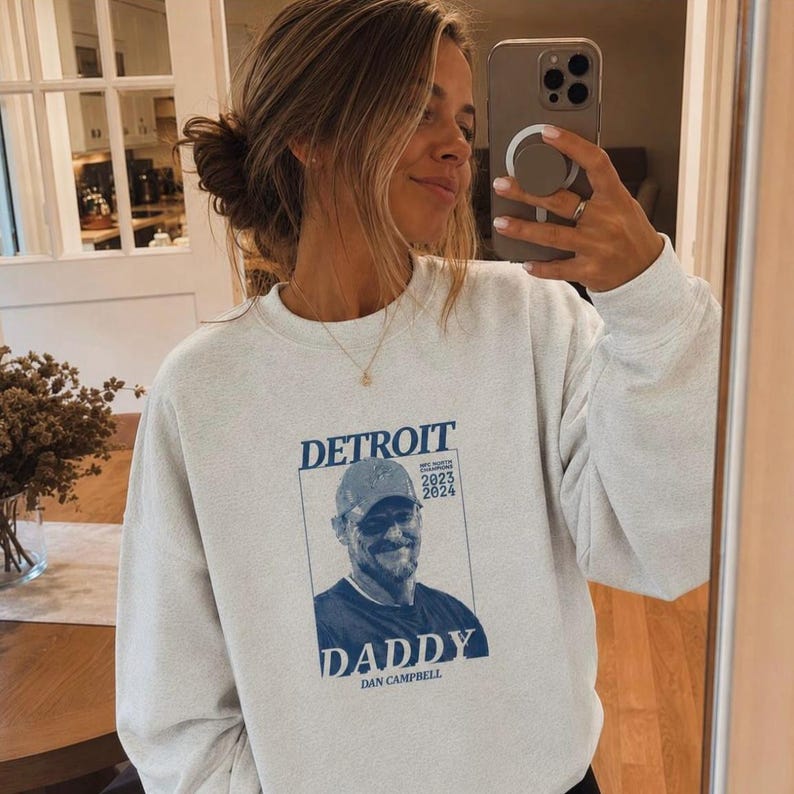 May include: A light gray crewneck sweatshirt featuring a blue graphic print on the chest. The design includes the text "DETROIT" at the top, "DADDY" at the bottom, "DAN CAMPBELL" below that, and "2023 2024" next to a portrait of a man wearing a baseball cap.