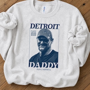May include: A light gray crewneck sweatshirt featuring a dark blue graphic print. The design includes a portrait of a man in a baseball cap, the text DETROIT, DADDY, DAN CAMPBELL, and NFC NORTH CHAMPIONS 2023 2024, all enclosed in a rectangular frame.