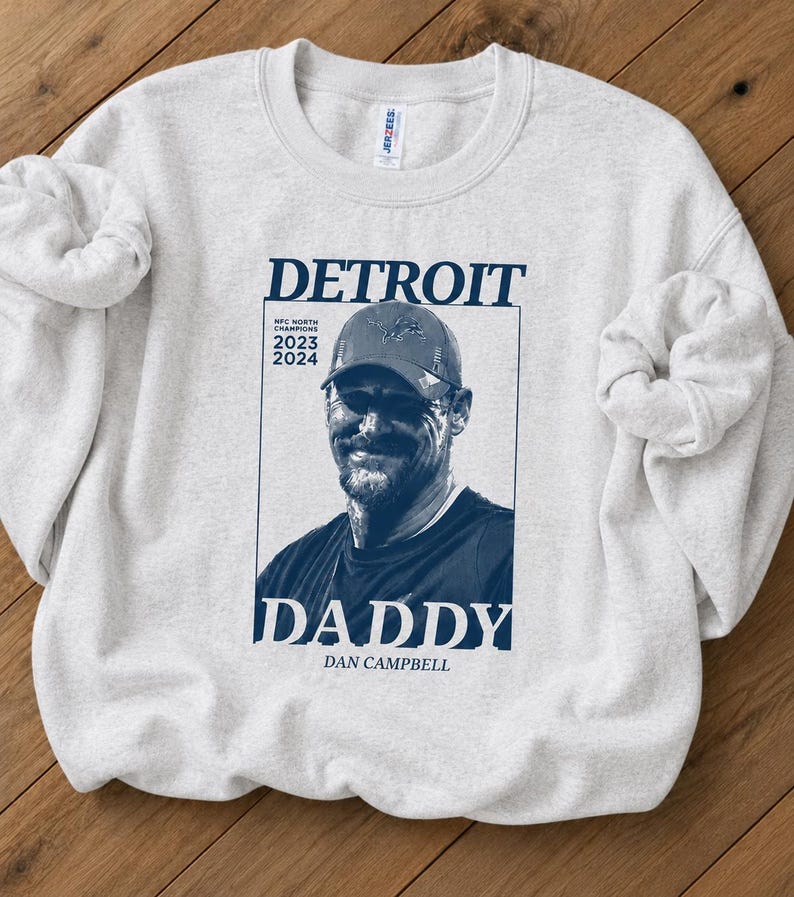 May include: A light gray crewneck sweatshirt featuring a dark blue graphic print. The design includes a portrait of a man in a baseball cap, the text DETROIT, DADDY, DAN CAMPBELL, and NFC NORTH CHAMPIONS 2023 2024, all enclosed in a rectangular frame.