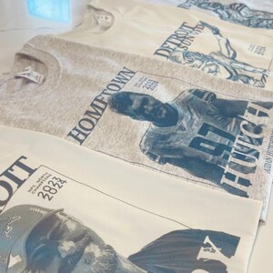 May include: Three crew neck t-shirts featuring graphic prints of football players. One heather gray shirt displays a player with the text HOMETOWN 97 HUTCH and NFC NORTH CHAMPIONS 2023 2024. Two cream shirts feature player graphics with DETROIT SUN GOD and NFC NORTH CHAMPIONS 2023 2024 text.