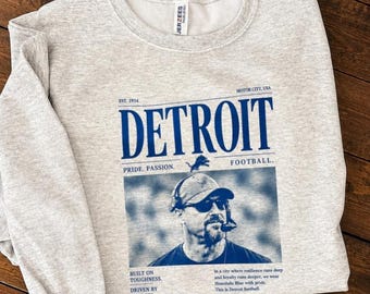 May include: A light gray heather crewneck sweatshirt featuring blue text and a graphic. The design includes the word DETROIT, the phrase PRIDE. PASSION. FOOTBALL., and a portrait of a person wearing a cap, sunglasses, and a headset microphone.